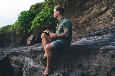 Gündüz vakti sakallı, dalgın ve kolları dövmeli bir erkek uçurumun yamacında oturuyor ve doğanın tadını çıkarırken deftere yazı yazıyor.