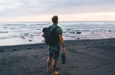 Tanımlanamayan bir erkek yolcunun kumsalda dalgalanan denizin yanında durup şişeyi ve sırt çantasını tutarken inanılmaz deniz manzarasının keyfini çıkarması.