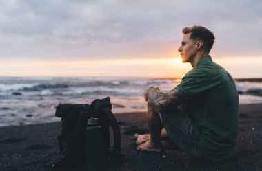 Gün batımında dalgalı denizle birlikte karanlık kumlu sahilde çantalı ve su şişeli rahat giysiler içinde genç bir erkeğin manzarası. Tatil sırasında gözlerini kaçırıp doğanın tadını çıkarıyor.