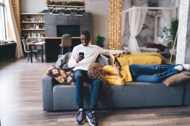 Orta Doğulu bir kadın kucağında uzanmış, modern bir oturma odasında telefonuna bakan, teknolojiyle meşgul rahat bir yaşam tarzı ve ev içi rahatlığı canlandıran siyahi bir adam oturuyor.