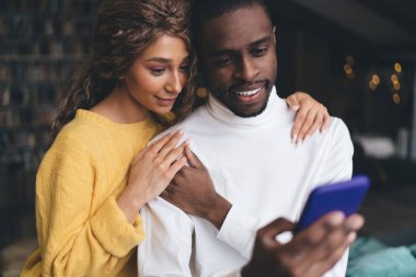 Sevecen ırklar arası bir çiftin yakın çekimi; sarı kazaklı beyaz kazaklı beyaz balıkçı yaka yakalı Afro-Amerikan bir adamı nazikçe tutan beyaz bir kadın, her ikisi de neşeli ışıkları olan evde telefona gülümsüyor.