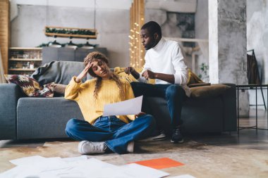 Genç bir Afro-Amerikalı erkek modern bir oturma odasında stresli bir beyaz kadınla finans tartışıyor, kağıtlar etrafa saçılmış, finansal planlama sorunlarını somutlaştırıyor.