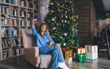 Mutlu, genç bir kadın, 30 'lu yaşlarının başında, kıvırcık saçlı, selfie çeken, aydınlatılmış ve süslenmiş Noel ağacının yanındaki koltukta oturmuş, hediyelerle çevrili, neşeli bir bayram havası yayıyor..