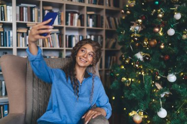 Mutlu, genç bir kadın, 30 'lu yaşlarının başında, kıvırcık saçlı, selfie çeken, aydınlatılmış ve süslenmiş Noel ağacının yanındaki koltukta oturmuş, hediyelerle çevrili, neşeli bir bayram havası yayıyor..