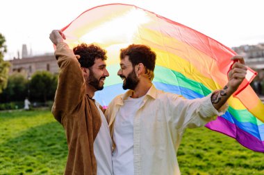 Mutlu genç eşcinsel çift LGBT bayrağı sallıyor ve çimenli çimlerde bulanık arka planda dikilirken birbirlerine bakıyorlar.