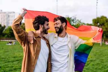 İspanyol sakallı, aşık homoseksüel erkekler gökkuşağı bayrağıyla yan yana durur ve yazın yeşil şehir parkının bulanık zemininde birbirlerine bakarlar.