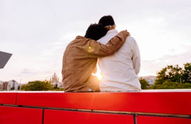 Genç erkek çiftin kırmızı taştan bir bankta otururken birbirine sarılışını doğanın bulanık arka planına ve romantik bir randevu sırasında kırsalda gün batımını seyredişini.