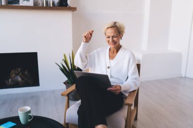 Orta yaşlı kadın serbest çalışan, elinde bir fincan sıcak içecekle masada oturuyor ve evdeki uzak projede çalışırken bilgisayarın ekranına bakıyor.