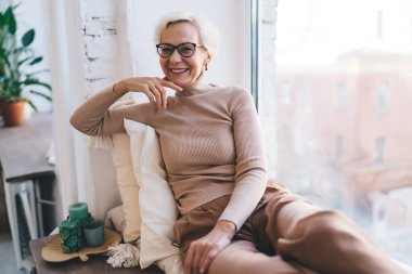 Gülümseyen, gri saçlı, günlük kıyafetler ve gözlüklü kadın emekliler pencere eşiğinde minderlere yaslanırken kameraya bakıyorlar.