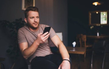 Günlük kıyafetlerle gezen, akıllı telefonlara bakan ve kafeteryada dinlenirken internetten bilgi arayan serbest sakallı bir erkek.