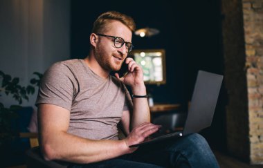 Gözlüklü bir erkek girişimci kafeteryada oturmuş bilgisayarla internetten ekranlara bakarken cep telefonuyla konuşuyor.