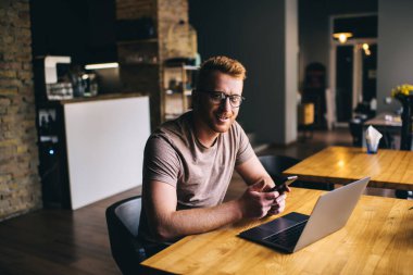 Modern kafeteryadaki gözlüklü, yüksek açılı, sakallı erkek serbest çalışan akıllı telefon sörfü yapan ve uzaktan bilgisayarla çalışan.