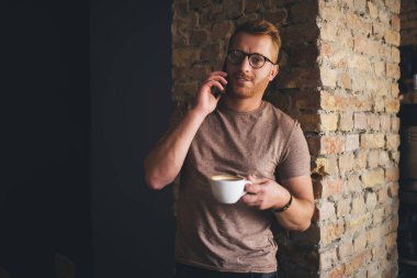 Kahve molası sırasında cep telefonu teknolojisi ile iletişim kurarken kameraya bakarak görüş düzeltmesi yapmak için optik gözlüklü yakışıklı sakallı adamın yarım boy portresi.