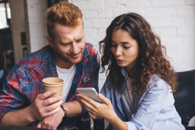 Birlikte zaman geçirirken, ahşap masada bir fincan kahve ve cep telefonuyla oturan, gündelik kıyafetli, pozitif genç bir çift.