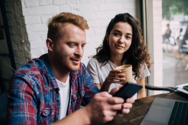 Pozitif genç iş arkadaşları dizüstü bilgisayarlarıyla masada oturuyor ve erkeklerin cep telefonlarını karıştırıyor ve elinde kahve fincanı ile kameraya bakarken bir yandan da kafede proje üzerinde çalışıyorlar.