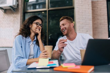 Pozitif genç erkek serbest yazar ve gözlüklü gülümseyen kadın dizüstü bilgisayarlı not defteriyle masada oturuyor ve kafede kahve içerken birbirleriyle konuşuyorlar.