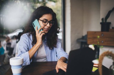 Neşeli, kıvırcık saçlı, hafif giyimli ve gözlüklü bir kız öğrenci internette internette internette kafede plastik kahve fincanıyla sohbet ederken sohbet ediyor.
