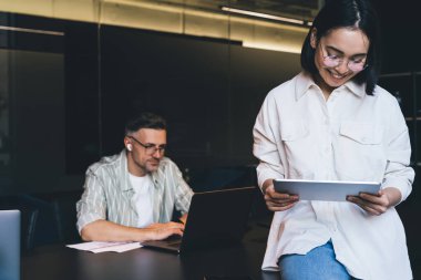 Görme düzeltmesi için mutlu Asyalı çalışan uygulama üzerinden internet sitesinde bilgi taraması için dijital tablet kullanıyor, neşeli grafik tasarımcısı dokunmatik ped ile online rezervasyon yapıyor