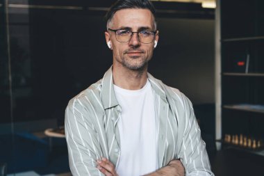 Beyaz tenli sıradan iş adamının yarı boy portresi görüş düzeltmesi için optik gözlüklü, kameraya bakan mavi kulaklıklı, elleri çaprazlanmış, yakışıklı, yetişkin bir erkek.