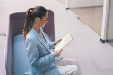 Yüksek açılı yan görüş, gündelik giysiler içinde genç bir bayanın parlak modern çalışma alanında çalışırken tablet üzerinde oturup notlar okuması.