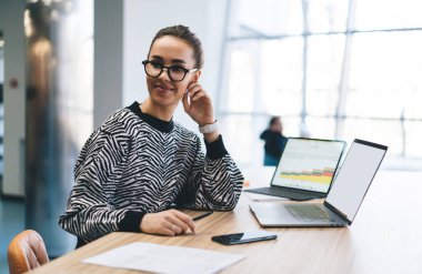 Gündelik giysiler ve gözlüklü genç kadın bilgisayar ve cep telefonuyla masada otururken modern çalışma alanında yazılım üzerinde çalışıyor.