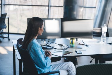 Modern çalışma alanı projesinde çalışırken masaüstü bilgisayarı ve cihazlarıyla masada oturan resmi kıyafet ve gözlüklü genç bayan programcının yan görüntüsü