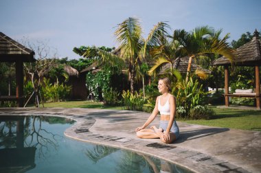 Sıska bir kadının, nilüfer çiçeğiyle havuz kenarında otururken, yeşil parkta yoga yaparken yüzünü çevirmesi ve tatil köyünde yaz tatilinin tadını çıkarması.