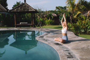 Namaste 'deki sıska bir kadının kolları havada, gözleri kapalı otururken havuz suyunun yanında bacak bacak bacak bacak üstüne atmış ve tropik tatil beldesinde yoga yaparken yan görüntüsü.