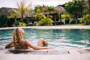 Yüzme havuzunda dinlenen genç sarışının arka görüntüsü. Elleriyle kenarına yaslanıp, güneş ışığında gözlerini kapayarak tatil köyünde tatilin tadını çıkarırken.