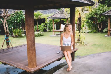 Sütunun yanındaki ahşap bankta dinlenen ve dizüstü bilgisayarlı akıllı telefonla oturup tropikal bahçede vakit geçiren çıplak ayaklı bir kadın vücudu.