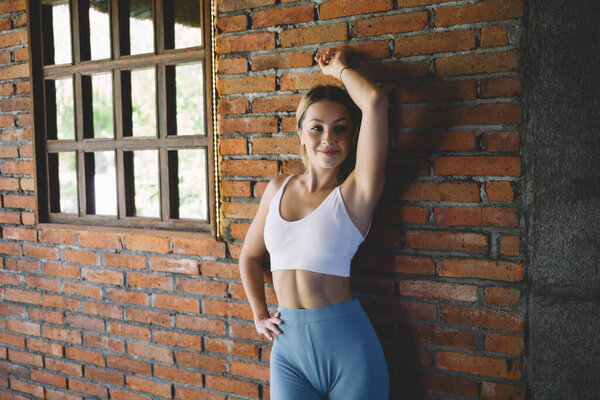 Happy young female looking at camera while leaning on back on brick wall under construction near window and stretching arm above head while touching waist with hand in daylight