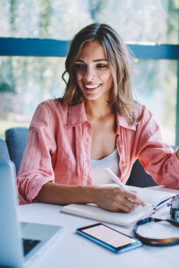 Başarılı iş webinar Web sitesinde izliyor ve notlar ile kablosuz internet kafede dizüstü bilgisayar kullanarak bir not defteri yapma mutlu genç kadın. Haber okuma ve yazma metin olumlu hipster öğrenci