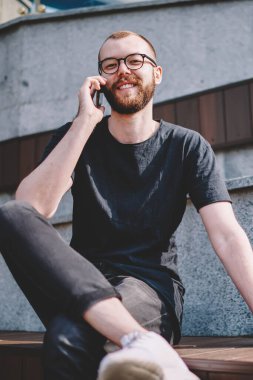 Gözlük smartphone aygıt üzerinde gezici arkadaşı ile iletişim sırasında kameraya gülümseyen sakallı neşeli genç adam portresi. Telefon aygıtı üzerinde arama olumlu hipster blogger