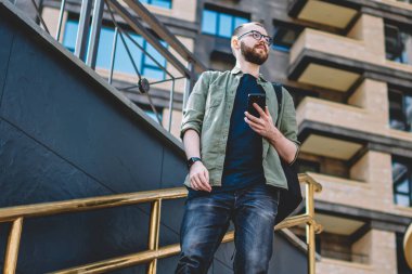 Durulması ayakta merdivenlerde smartphone ile kentsel ortamda eller iken uzakta görünen genç adam sakallı. Hücresel cihaz tutan yolda rahat kıyafet düşüncesinde düşünceli hipster adam giyinmiş
