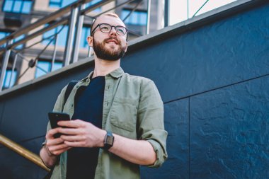 Aşağıda görünümü düşünceli erkek blogger düşünce modern smartphone kentsel ortamda merdivenlerde açık havada duran üzerinde internet yayıncılığı. Düşünceli hipster adam uzakta süre irsal sms arıyorsunuz