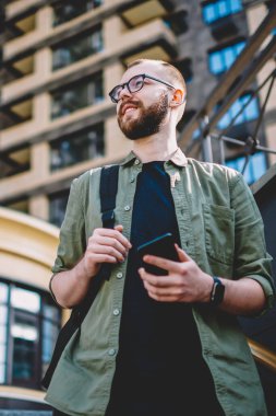 Aşağıda görünümü durulması sakallı öğrencinin elinde smartphone ile yürüyüş sırasında şehir zevk. Düşünceli hipster adam uzağa bakarak ve modern telefon tutarak gülümseyen gözlük rahat giyinmiş
