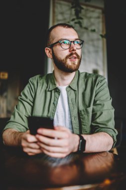 Düşünceli uzağa bakarak ve internet yayıncılığı modern smartphone aygıtında web sitesinde yazmak için fikirler üzerinde düşünme erkek blogger sakallı. Sosyal ağlarda profil güncelleştirme durulması hipster adam