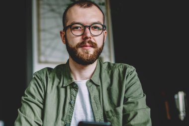 Kafede ücretsiz Wi-Fi kullanarak modern smartphone içinde online sohbet ederken kameraya bakarak gözlük sakallı genç adam portresi. Hipster blogger cep internet bağlantısı üzerinden App yükleme