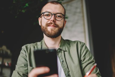 Modern smartphone elinde tutan ve kameraya ararken internet sitesi bilgileri yeniden yazma vizyon düzeltme için şık gözlük sakallı hippi öğrenci portresi