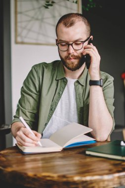 Smartphone üzerinde arama ve Not Defteri'nde kafede kahve masada oturan aşağı yazma dalgın sakallı genç adam. Ciddi hipster adam cep telefonu konuşma sırasında not defterinde kayıt yapma
