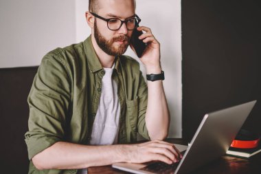 Dalgın smartphone aygıtında çağrılırken dizüstü bilgisayarda posta kontrol uzmanı. Sırasında cep telefonuyla konuşan genç adam sakallı kablosuz internet kullanarak modern ağ dizüstü bilgisayarı, uzaktan çalışma