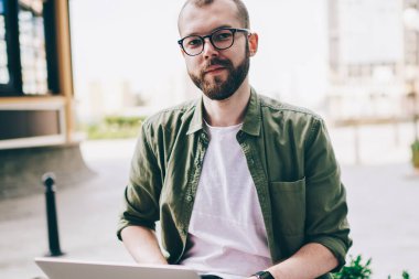 Serbest, genel 4 g kullanarak dizüstü bilgisayar üzerinde çalışırken kameraya bakarak gözlük sakallı genç adam portresi Internet. Mesafe işi sırasında netbook ile sokakta oturan hipster adam