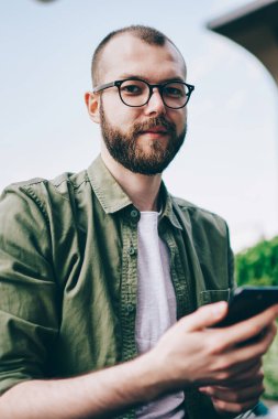 Telefon elinde tutan ve uygulaması Internet bağlantısı kullanarak güncelleştirme gözlük sakallı genç adam portresi. Casual giyinen hipster adam kameraya bakarken smartphone programı yükleme
