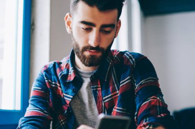 Ciddi genç hipster adam kampüste otururken akıllı telefonuyla para transferi yapıyor. Milenyum erkeği sosyal ağlarda cep telefonu haberlerini içeride kablosuz internet üzerinden kullanıyor.