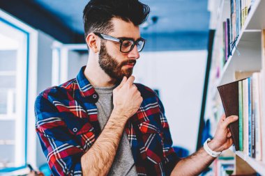 Düşünceli erkek kitap kütüphane üniversite duran raf yakınındaki seçme, hipster adam rahat giyinmiş malzeme çekme edebiyat konsantre ve favori kitapçıda araştırma yapma gözlükler