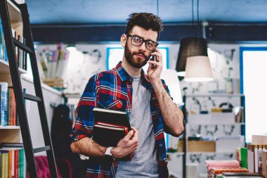 Satın almak, üniversite kitaplığında vakit şık genç adam için müşteri hizmetleri çağrı ve kitap seçerken kredi kartı üzerinde denge kontrol yakışıklı bin yıllık erkek öğrenci portresi