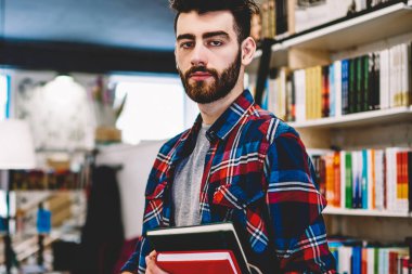 Yarım uzunluk portre zeki yakışıklı erkek genç üniversite kütüphanesinin rafları kitaplarla, zeki öğrenci bilgi ve belgeleri arama yaparken kameraya bakarak serbest zaman geçirmek