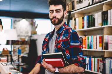 Yarım uzunluk portre zeki yakışıklı erkek genç üniversite kütüphanesinin rafları kitaplarla, zeki öğrenci bilgi ve belgeleri arama yaparken kameraya bakarak serbest zaman geçirmek