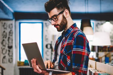 Üniversite kütüphanesinde kitaplarla, teknoloji ve iletişim konseptiyle rafların yanında dururken çevrimiçi iş için bilgisayar güncelleme yazılımında yazı yazan profesyonel bir erkek.