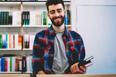 Gülümseyen, yarım uzunluk portre raf, edebiyat seçmek ve araştırma kitapçıda kazanmak mutlu başarılı hipster adam ayakta Üniversite Kütüphanesi kitap seçme erkek genç sakallı